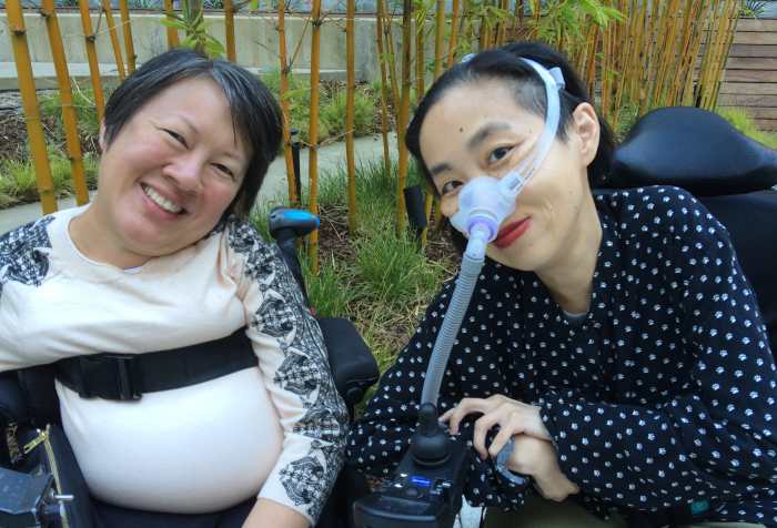 On the left is Ing Wong-Ward, a Chinese Canadian disabled woman with short black hair and a cream top with black decorative print on the sleeves. She is in a wheelchair with a belt across her chest. On the right is Alice Wong, A Chinese American disabled woman with short black hair and a black shirt with small white cat paw prints. She is in a wheelchair and wearing a mask over her nose attached to a tube for her ventilator.