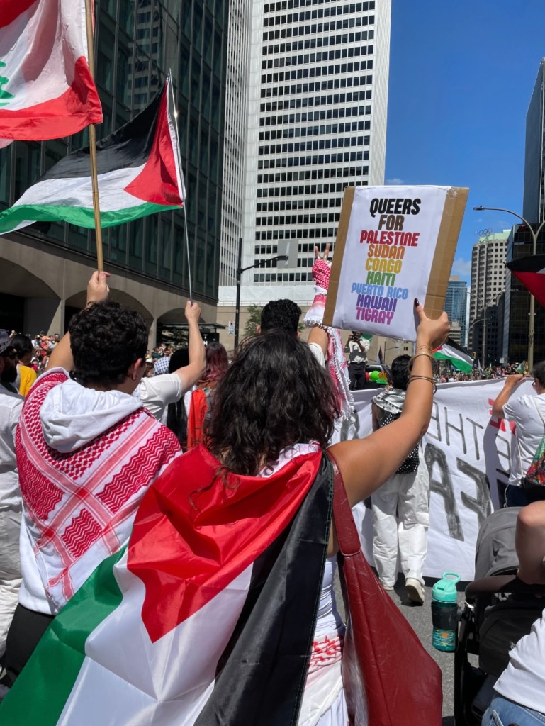 Street protest in Montreal, in a setting surrounded by tall modern skyscrapers with glass and steel facades. The sky is clear and blue, indicating a sunny day