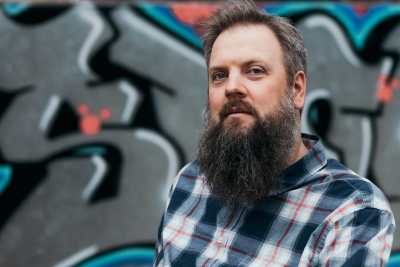 A white male about 40 years old with a long beard and greying hair looks at the camera. He wears a blue and red plaid shirt. Behind him is a mural silver, pink, and blue graffiti lettering.
