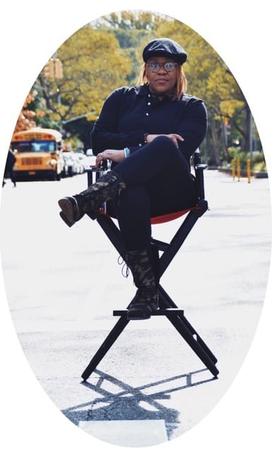 Oval photo of Jade Bryan, a Black Deaf woman sitting in a director’s chair outdoors with trees and parked cars behind her. She is wearing all black, and a black beret and glasses. Her left leg is crossed over her right, both arms folded.