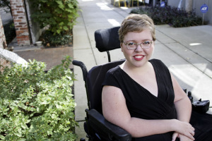 White woman with short blonde hair with glasses. She is wearing a black v-neck top and sitting in a wheelchair outdoor next to some bushes.