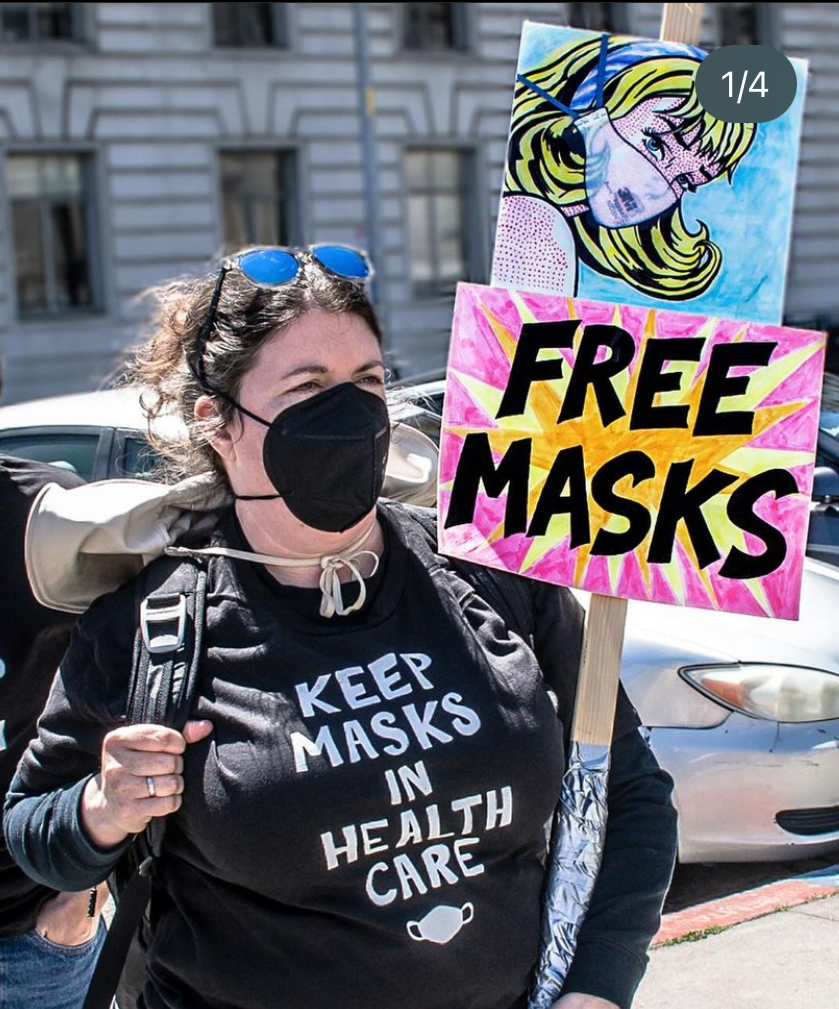 A photo of Britta Shoot, a white woman with brown hair wearing a black respirator mask and sunglasses on her head. She is holding a sign that reads FREE MASKS and wearing a black t-shirt that says in white letters KEEP MASKS IN HEALTH CARE