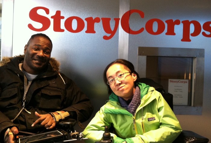 2 individuals in wheelchairs in front of a StoryCorps listening booth. The person on the left is a middle-aged African American man in a black jacket w/ short braids. The person on the right is a middle-aged Asian American woman wearing a green jacket and glasses. Both are smiling at the camera.