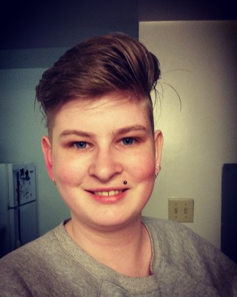 Image Description: A head and shoulders shot of a white smiling woman in her early 30s wearing a grey sweater. She has short blond hair styled in an undercut.