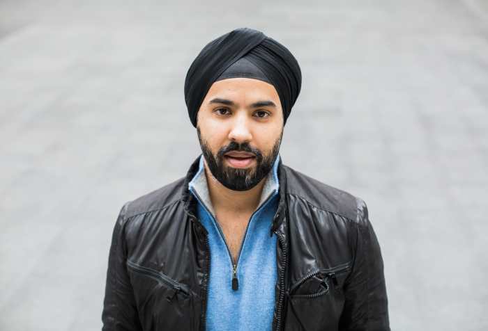 Sikh man wearing a black turban, a black leather jacket and a light blue shirt underneath. He has a beard with black hair and standing outdoors with a gray background. Photo credit: Les Talusan