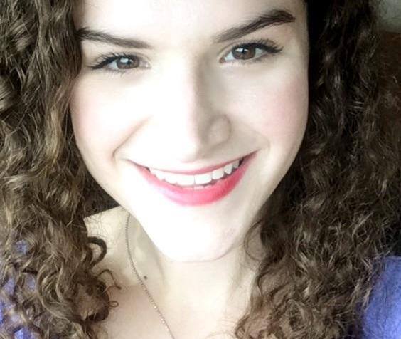 A photo of Lillie Lainoff taken in July 2016. She appears to be white and is wearing a purple shirt and a sea turtle necklace. She has long, curly brown hair and is smiling at the camera.