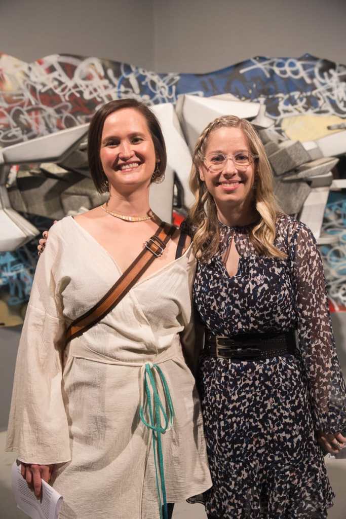 Photo of curators Vanessa Chang and Lindsey D. Felt standing shoulder to shoulder in front of a graffiti installation, beaming at the camera. Lindsey has wavy blonde hair and wears translucent glasses and a gauzy black and white dress with a black double buckle belt. Vanessa has a short dark brown bob and wears a gold choker necklace and a long sleeved white kimono top. A brown bag strap crosses her chest.