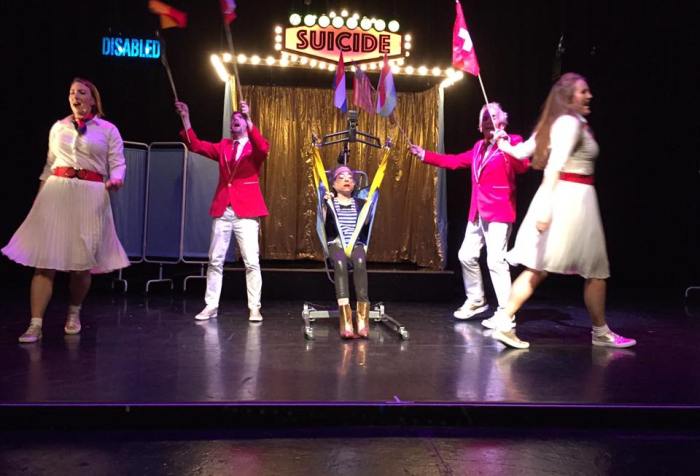 Photo of stage performance of "Assisted Suicide, the Musical" The main character, a thin, petite white woman is Liz is seated in a hoyer lift on centre stage. There is gold curtain behind her with a sign 'suicide' in lights. Two women wearing white tops and skirts with red belts are dancing, with two men in red jackets with white trousers are holding up flags.