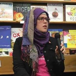 Image description: "A white woman with purple eyeglasses sitting down in front of a wall of books holding a microphone. She is wearing a black denim jacket over a pink shirt, and a purple hijab with broad patterned yellow, blue and pink stripes falling over her shoulder"