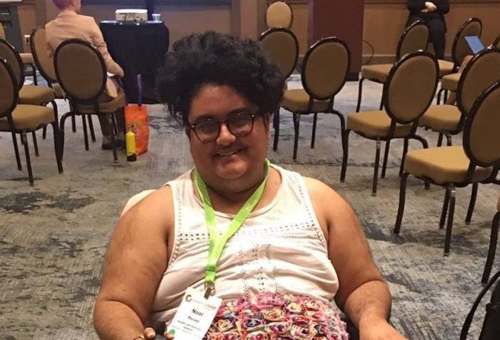 A south asian person poses in a Jazzy Passport folding power chair. He has on brown lace up boots, a colorful floral skirt, and a white lace top. He has a conference badge. He is smiling.