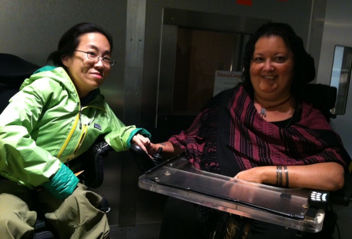 two women of color in wheelchairs in front of a recording booth. The woman on the left is wearing a green jacket, glasses, and has black hair. The woman on the right has long curly dark hair, a black and magenta top and a tray attached to her wheelchair. Both are smiling.