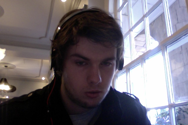 Image of a white man with brown hair staring at the camera. He is wearing headphones.