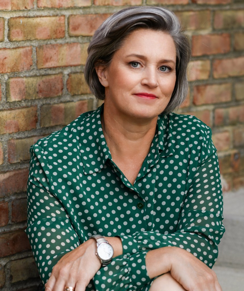 Margaret Price, a white genderqueer femme, wears a green polka-dot dress and sits in front of a brick wall. Her chin-length hair is gray and her face is smiling slightly.