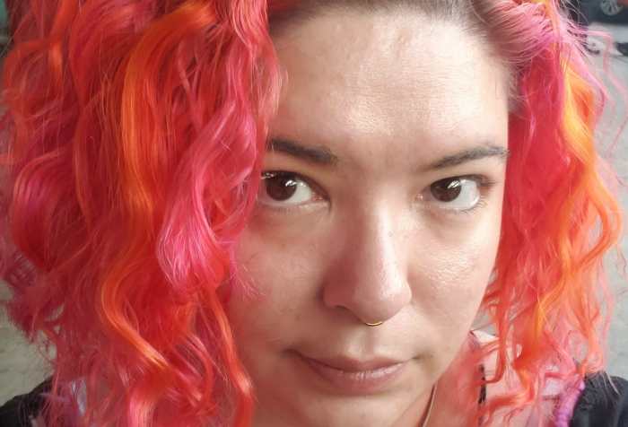 Headshot of a Light skinned indigenous non-binary person with short bright pink and orange hair. She has brown eyes and a septum piercing. She is wearing a pink striped shirt with a black cardigan over top. She has two heart tattoos on her collar bones.