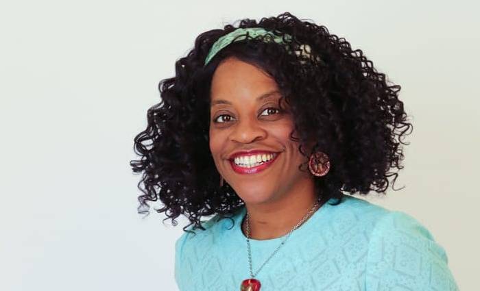 Robin, a brown woman, is posed and smiling. Wearing a light blue-green lace vintage dress, she is holding the handles of her walker.