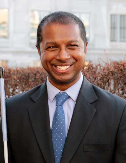 Picture of Sachin Pavithran, a South Asian man in a suit holding white cane outside the Capitol in Salt Lake City.