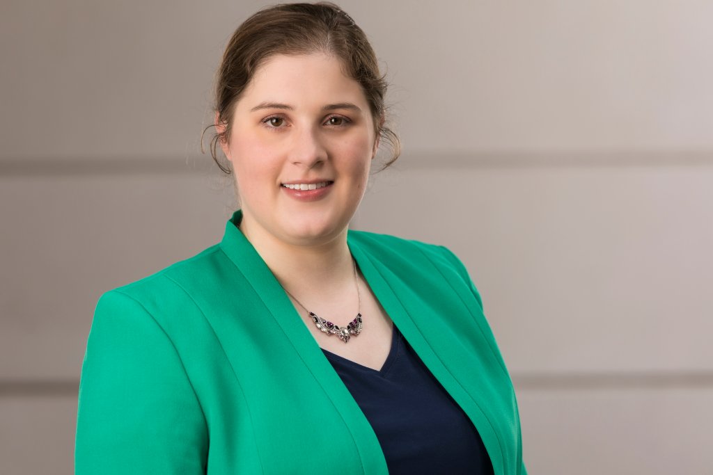 Sarah, a white woman with brown hair pulled back, smiling at the camera. She is wearing a green blazer and navy blouse.