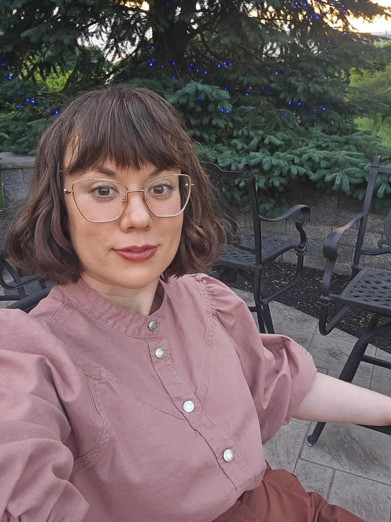A young white woman sits outdoors, smiling at the camera. Her brown hair is cut in a long bob with bangs and she has oversized rose-gold glasses. She wears a pink blouse with silver buttons down the front and brown slacks. In the background you can see a patio with iron chairs, and a fir tree lit up with blue lights for the holidays.