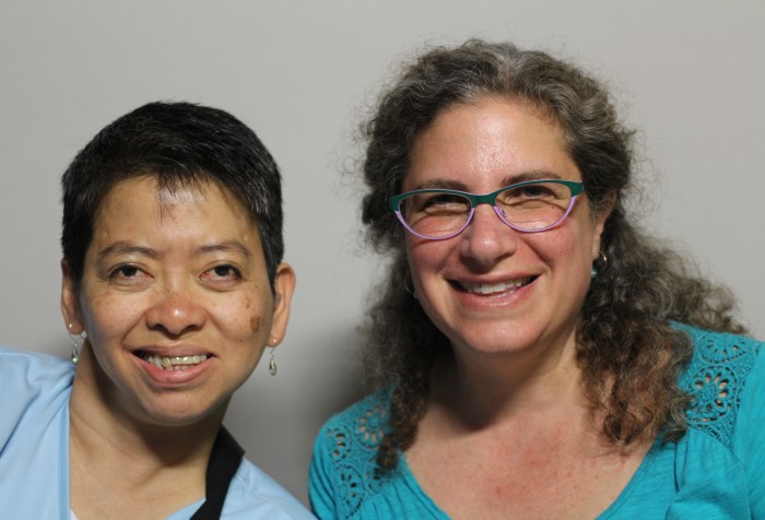 Image Description: A photo featuring Suzanne Levine and Yvette Fang taken on October 4th, 2014. The woman on the left is Yvette Fang. She appears to be of Asian descent, has short dark hair, is looking at the camera and smiling. She is wearing earrings and a light blue shirt. The woman on the right is Suzanne Levine. She appears to be white, has long curly brown hair that is pulled back in a half ponytail and is wearing glasses and an aqua colored shirt. She is looking at the camera and smiling.