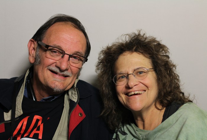 A photo featuring Tom Olin and Marilyn Golden taken on November 19th, 2014. They are both looking at the camera and smiling. The person on the left is Tom Olin. He appears to be white, and is wearing a black jacket and a black t-shirt. He has dark hair combed to the side and is wearing glasses. The person on the right is Marilyn Golden. She appears to be white, and is wearing a light green shirt. She has long brown hair and glasses.