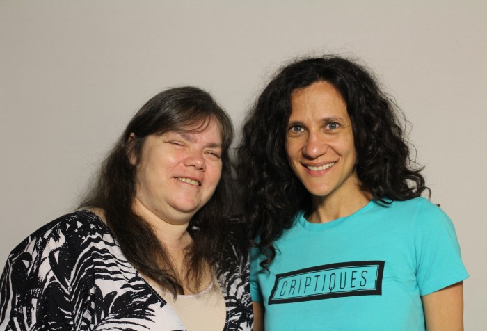 A photo featuring Lavaun Heaster and Cheryl Green taken on April 22, 2015. The woman on the left is Lavaun Heaster. Her family is Hispanic, Native American and white, and she appears to be white. She is wearing a black and white cardigan with a beige tank top. Her hair is long and brown, and she is smiling at the camera. The woman on the right is Cheryl Green. She appears to be white, has long, wavy, dark brown hair, green eyes and is smiling at the camera. She is wearing a turquoise tee-shirt that says the word “criptiques” printed in a black-framed rectangle.