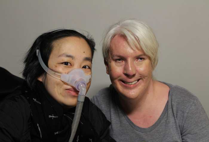 On the left, Alice Wong an Asian American woman wearing a black jacket and black scarf with little white daggers. She is wearing a mask over her nose attached to a gray tube which is connected to her ventilator. On the right is Jennifer Justice, young white woman with very light long blonde hair pulled back. She is wearing a heather gray scoopneck shirt. She is smiling at the camera.