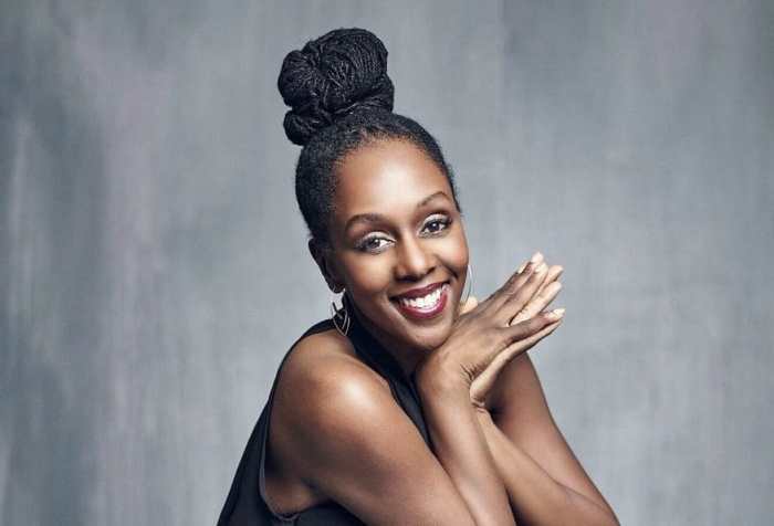 Stephanie Thomas, a Black woman with oval shaped eyes, round nose, smiling. Her chin is resting on her clasped hands. Her hair locs are pulled up in a high bun and wearing a sleeveless black top. The background is a grayish black backdrop. My condition’s name is Symbrachydactyly. I am a congenital amputee missing digits on my right hand and feet. Photo credit: Benjo Arwas @benjoarwas