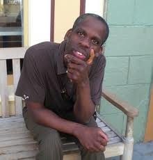A black man sitting at a wooden bench and one hand is raised, at his chin. He is wearing brown slacks and a brown t-shirt. He is staring right at the camera.