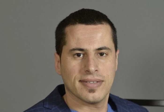 Photo of Mustafa Rfat, a man with short dark brown hair wearing a navy blue blazer with v-neck blue shirt. Behind him is a dark gray background.