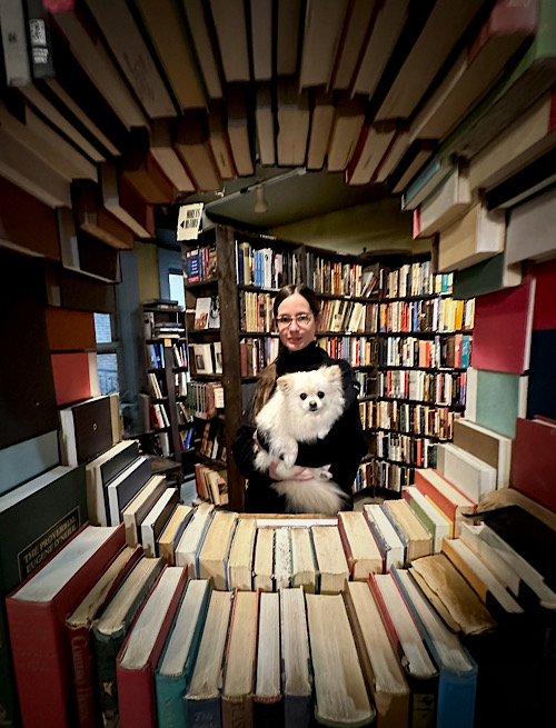 A white woman wearing glasses and a black sweater smiles while holding a small, fluffy white dog in her arms. They are surrounded by tall shelves filled with books, and are gazing together at the camera through a circular “window” opening in a sculptural wall made of hardcover books in an array of colors.
