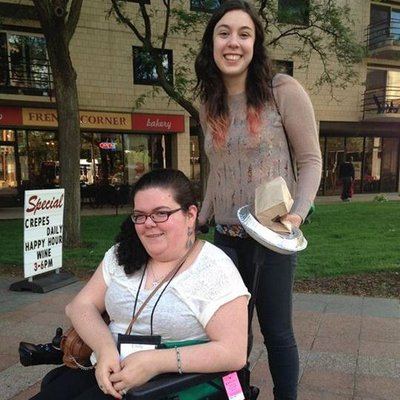 Two young women smiling at the camera. One has brown hair in a ponytail and glasses, she is sitting in a wheelchair. Another young woman is standing behind her with shoulder-length brown hair.