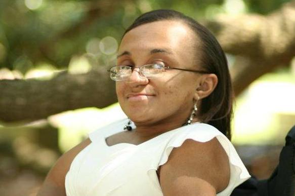 Photo of a young Black woman with her hair parted and hanging down straight. She is smiling and facing the camera sideways in her wheelchair. She is outside under the tree branches while wearing a white and black dress.