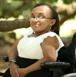 Image description: Photo of a young Black woman with her hair parted and hanging down straight. She is smiling and facing the camera sideways in her wheelchair. She is outside under the tree branches while wearing a white and black dress.