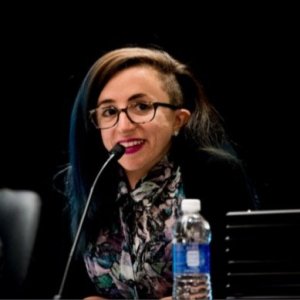 A young white woman wearing eyeglasses with long hair with blue streaks. She is looking sideways and smiling with a microphone in front of her. She is wearing a multicolored print shirt with a black jacket. The background is black.