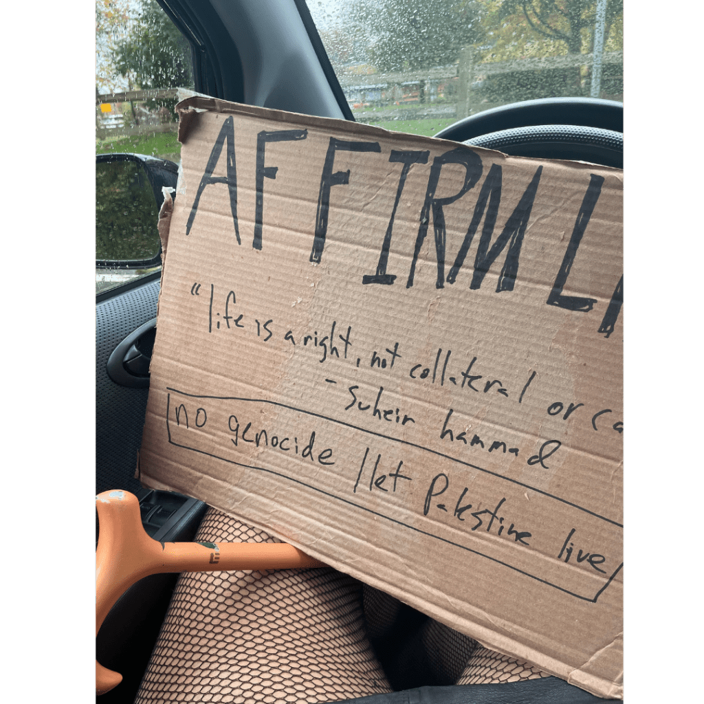 a close up of Leah's sign in their car, it is made of brown cardboard and reads AFFIRM LIFE and underneath "life is a right, not collateral or casual"- Suheir Hammad and the words "no genocide/ let Palestine live" are visible. The sign rests on Leah's knees in fishnets, their cane is visible stuck to the left of where they are sitting in the driver's seat, behind their steering wheel.