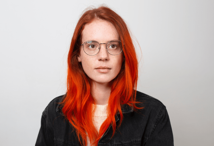Color photograph of Beatrice Adler-Bolton, simple portrait photo of a pale, freckled white woman with natural red to dyed orange long wavy hair, wearing round gold rimmed glasses and a black t-shirt in front of a white background.