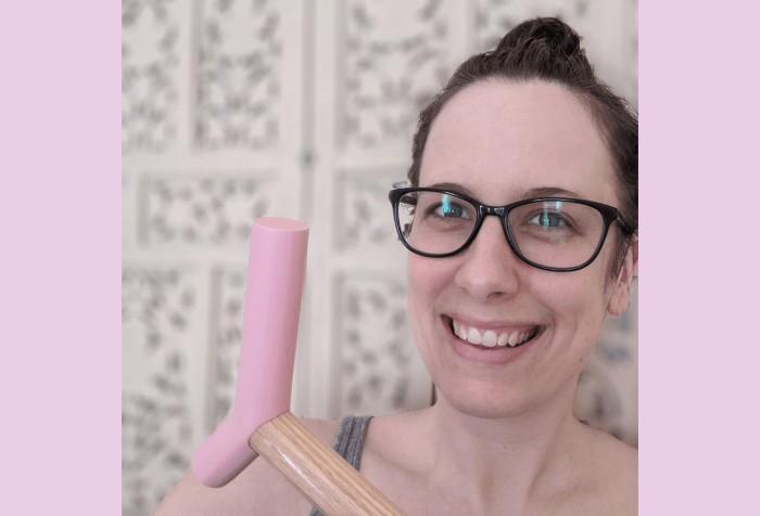 Graphic with a soft pink background featuring a photo of Brianne, a white woman in her mid-30s wearing black framed glasses and dark brown hair in a messy bun, smiles at the camera. She’s wearing a grey tank top, holding the pink epoxy handle of her blonde wood cane up beside her face and sitting in front of a blurred off-white decorative screen. The photo has been cropped close around Brianne’s face, shoulders and cane handle.