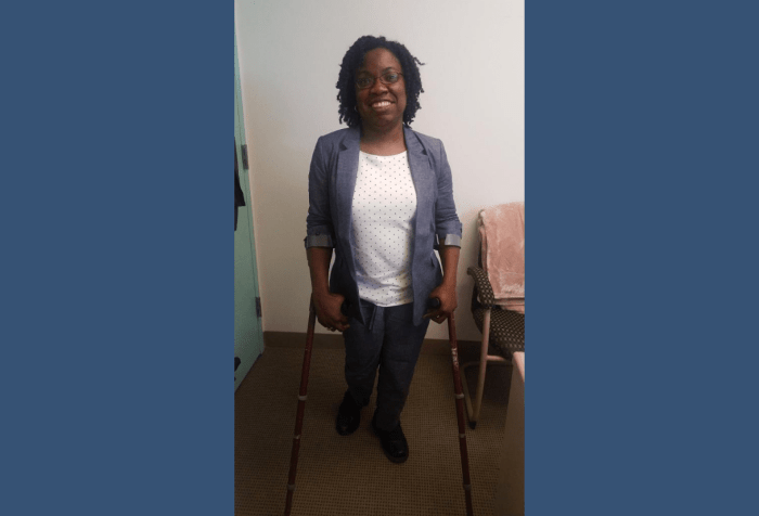Graphic with a dark blue background with a photo of Britney Wilson, Black woman with curly natural, black hair and glasses in a blue chambray suit and white and blue polka dot top standing on crutches in an office.