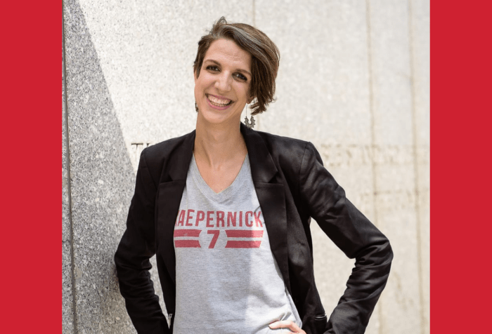 Graphic with a dark red background and in the center a photo of a white woman, smiling and standing with her hands on her hips and head slightly tilted to the left, in front of a gray granite wall. She has short brown hair in an asymmetrical haircut, longer on her left side and shorter on her right side. She is wearing dangling silver earrings and a gray t-shirt that reads "Kaepernick 7" in red, underneath a black blazer. 