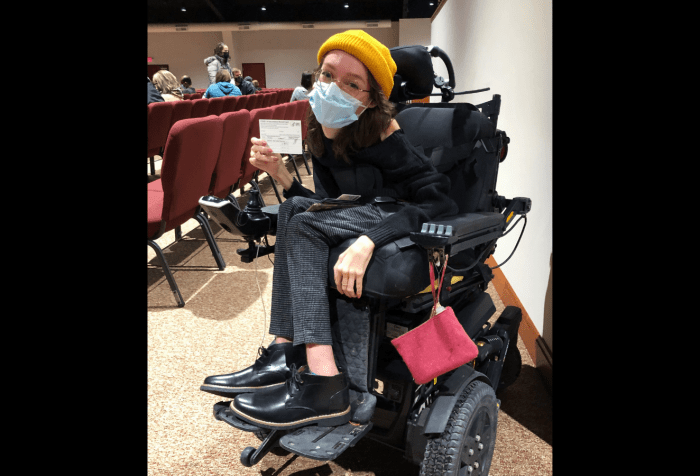 Graphic with a black background and a photo of Emily Ackerman, a white woman in a black power wheelchair is holding up her COVID vaccine card. She’s wearing a black sweater and boots with grey checked pants, a yellow beanie, and a blue medical mask. 