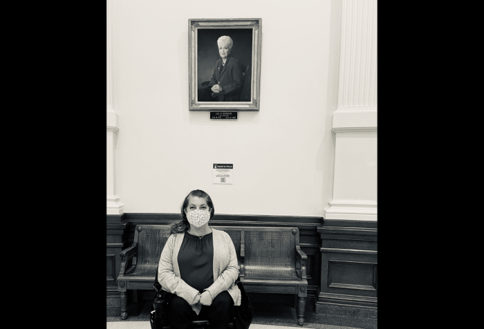 Graphic with a black background and in the center is a black and white photo of Emily Wolinsky sitting in her power wheelchair in front of a portrait of the late Governor of Texas, Ann Richards. 
