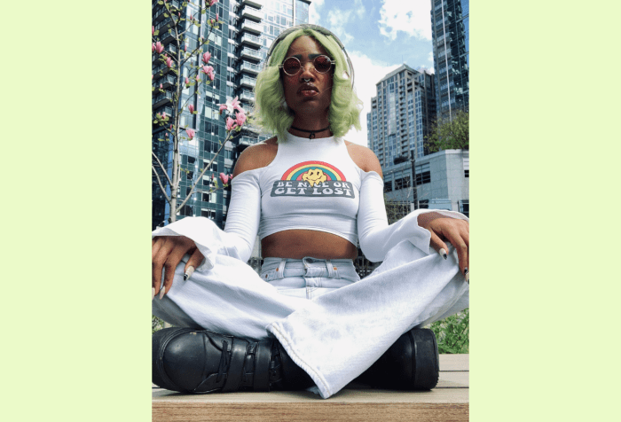Image of Faati, a nonbinary light brown skinned Black Native, sitting cross legged in front of a cherry blossom tree and multiple high rise buildings. They have on acid washed light blue bell-bottom jeans, a white crop topped long sleeved shirt and black platform boots. They are sitting cross-legged with their hands on their knees. Their fingernails are a long almond acrylic style with nude coloring and black and white designs on top. They have green straight 70s styled hair with the ends bumped. They are wearing circle 70s style red lens sunglasses.