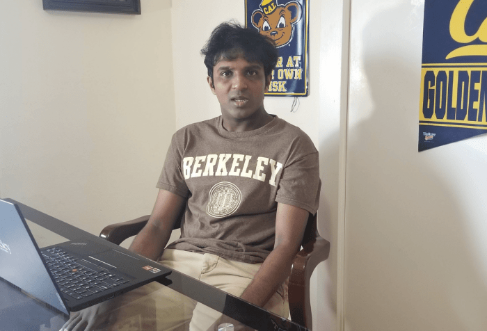 The image shows a young American male in his 20s of Indian American descent, with brown skin and black hair. He is sitting at a desk with a laptop open in front of him with some books on the side of the table. The laptop has the word Berkeley on it as does the brown t-shirt he is wearing. Behind him on the wall are college banners and posters with words like Cal, Golden Bears, and catchy phrases like, Bear Den, enter at your own risk.