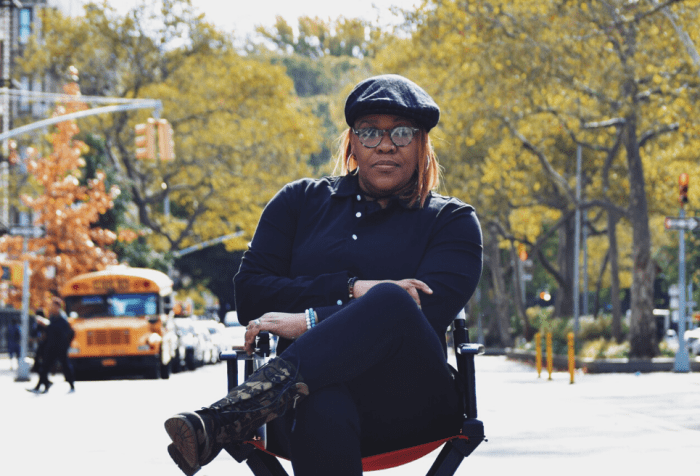 photo of Jade Bryan, a Black Deaf woman sitting in a director’s chair outdoors with trees and parked cars behind her. She is wearing all black, and a black beret and glasses. Her left leg is crossed over her right, both arms folded.