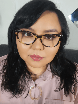 Picture of Johnnie Jae, black hair, bangs brushed to the right side of her face. She is wearing glasses that are slightly too big for her head, nude pink lipstick, a dusky pink shirt with a necklace featuring a round citrine pendant.