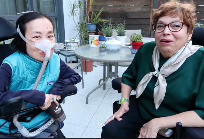 An outdoor patio with Alice Wong [left], an Asian American disabled woman in a wheelchair. She is wearing a mask over her nose connected to a tubing for her ventilator. She is wearing a long-sleeved navy shirt and a sporty aqua blue vest and holding a paper coffee cup. On the right is Judith Heumann, an older white woman with short wavy brown hair. She is sitting in a power chai and wearing glasses, a dark green short-sleeved top and a scarf tied into a knot. Both are looking at the camera and smiling.