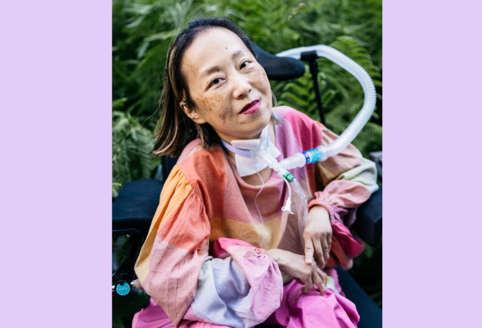 Graphic with a purple background. In the center is a photo of an Asian American woman in a wheelchair with a tracheostomy at her neck that has a tube connected to her ventilator. Her head is tilted to the left side, she is smiling, and she is wearing a magenta lip color. Her shirt is plaid with pale pink and other pastel colors and she is wearing pink pants. Behind her is a lot of greenery from bushes and trees at a park. Photo by @allisonbusch_photography