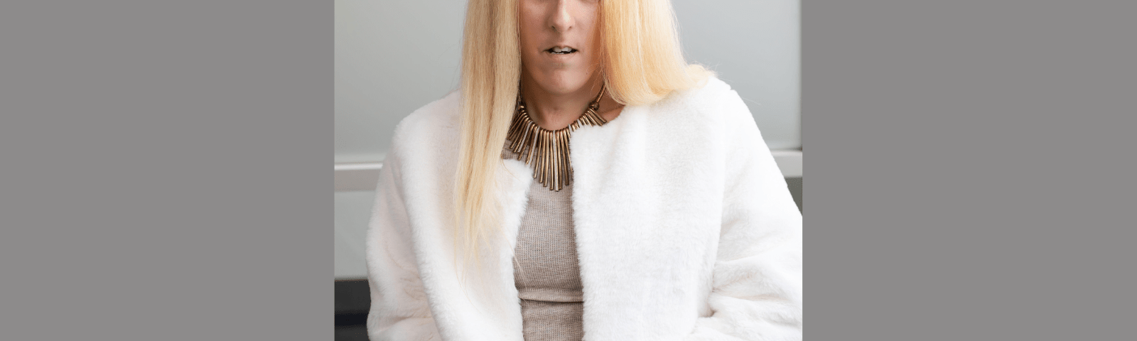 A headshot of a woman with brown color eyes and olive-tan skin complexion with long, straight blonde hair, wearing a baige top with a white jacket and a gold necklace. 