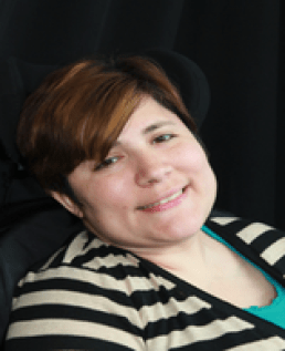 Black background of a light skinned woman with brown hair with gold streaks, brown eyes, sitting in her wheelchair wearing an aqua green undershirt and a beige and black striped sweater. 