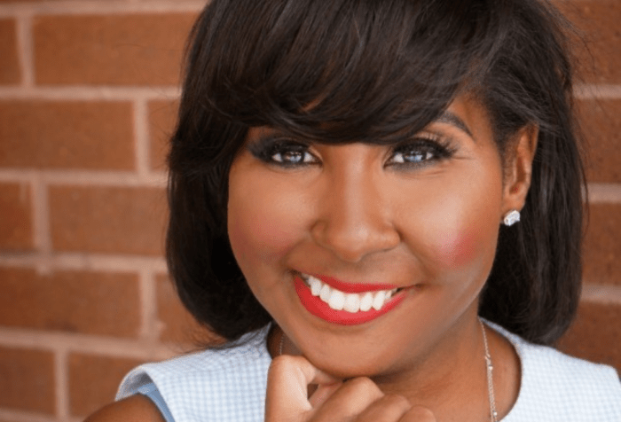 Rasheera Dopson, MPH, a Black woman with medium-dark brown hair. She is wearing a sleeveless blue and white checkered top, with a silver necklace, wearing red lipstick and smiling.